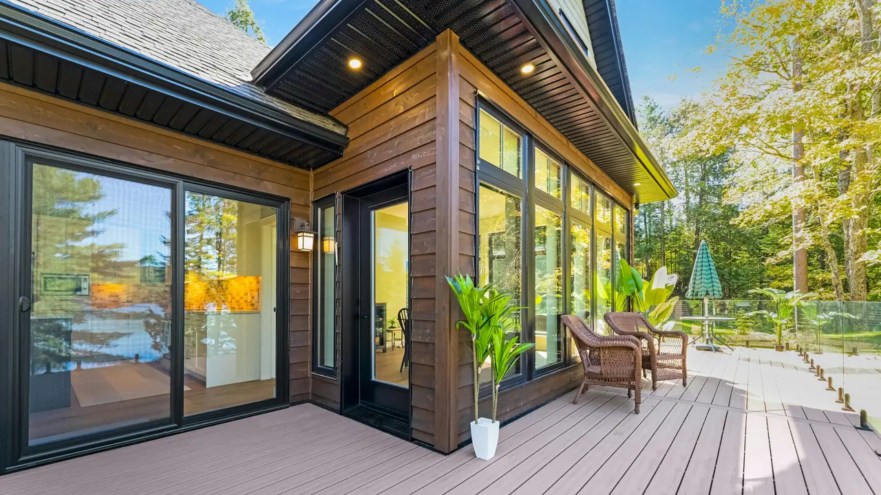 Modern home with a wide composite deck, glass railing and wicker chairs beside tall black-framed windows