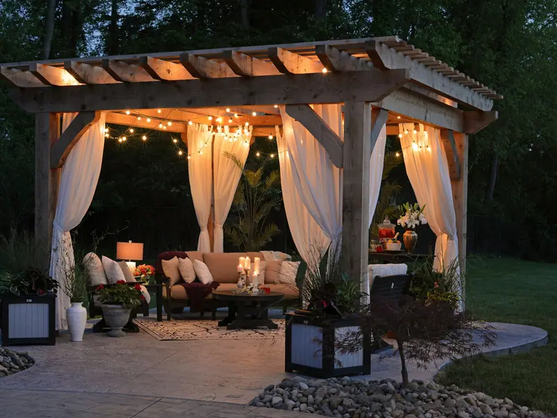 Garden pergola with white curtains, string lights and lounge seating at dusk
